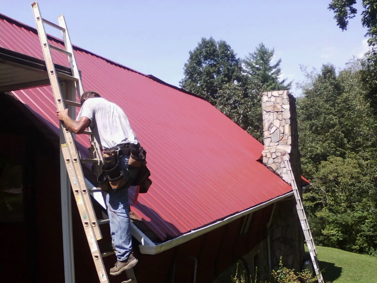 Roofer on a ladder beside a red metal roof and stone chimney — Soffit & Fascia Repair by Roofing Brothers in Wellfleet